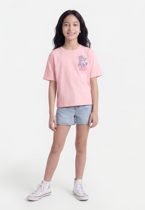 Νεαρή κοπέλα όρθια, φοράει ροζ μπλούζα Care Bears, ανοιχτόχρωμα τζιν σορτς και λευκά ψηλά αθλητικά παπούτσια μπροστά σε λευκό φόντο.