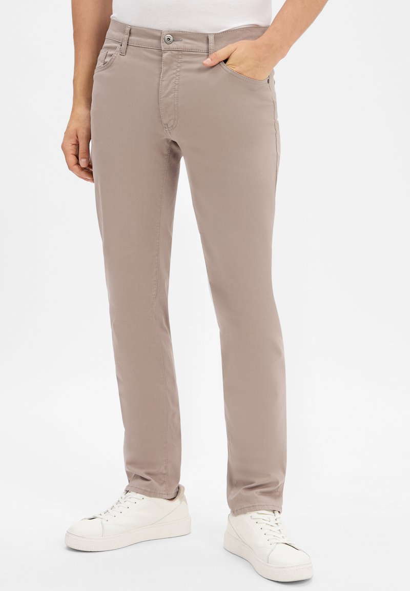 Osoba ubrana w beżowe, slim-fit spodnie z rękami w kieszeniach i białe, casualowe sneakersy, stojąca na białym tle.