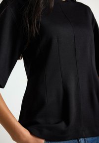 Top nero a maniche corte realizzato in tessuto liscio, con scollo tondo e dettagli sottili a pannelli sul davanti. Design minimalista.