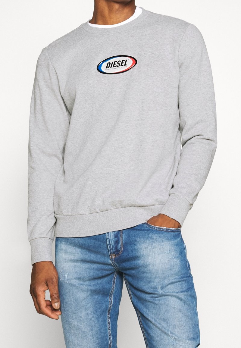 Homme portant un sweat-shirt Diesel gris clair avec logo sur la poitrine et un jean bleu, main dans la poche, debout devant un fond uni.