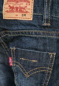 Jeans Levi's de denim azul oscuro, con costura amarilla, una etiqueta de cuero con texto rojo, y un pequeño bolsillo trasero con diseño de costura curvada.