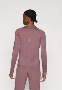 Αθλητικό τοπ με μακριά μανίκια σε μουβ ύφασμα. Διαθέτει ραγκλάν μανίκια, στρογγυλή λαιμόκοψη και σχισμές αερισμού στην πλάτη. Λείο υφή.