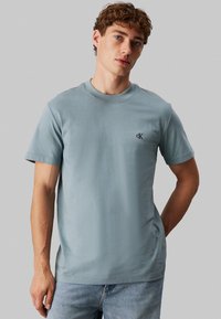 Camiseta de algodón azul claro con cuello redondo, mangas cortas y el logo "ck" en el lado izquierdo del pecho. Diseño sencillo con una textura suave.