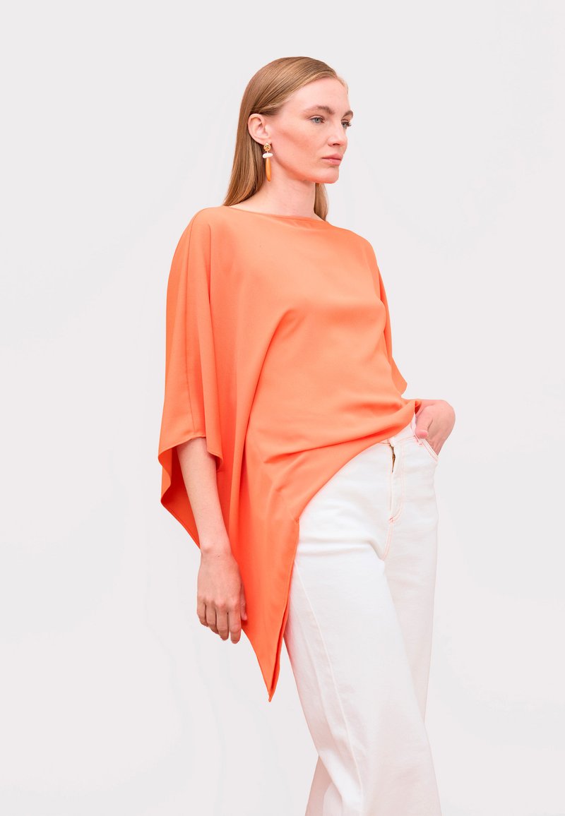 Žena v voľnom oranžovom topu a bielych nohaviciach s vysokým pásom, pózujúca s jednou rukou v vrecku na jednoliatom pozadí.