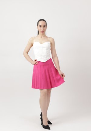 Mujer de pie con corsé blanco, falda plisada rosa y tacones altos negros sobre un fondo blanco liso.