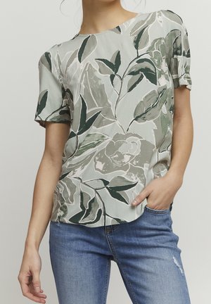 Persoon die een kortemoublouse draagt met grote groene en grijze bladpatronen, gecombineerd met blauwe versleten spijkerbroek, hand in de zak.