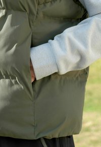 Olivgrüne, gepolsterte Weste mit einer Seitentasche. Hellgrauer, gerippter Bund sichtbar, aus weichem Stoff. Glatte Textur, lässiges Design.