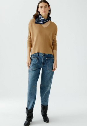 Beige gebreide trui met V-hals, gecombineerd met blauwe denim jeans met zakken en een relaxte pasvorm. Zwarte kniehoge laarzen en een marineblauwe sjaal.