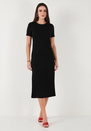 Black, short-sleeved, form-fitting dress with a knee-length hem and side slits; smooth fabric; worn with red sandals.
