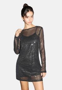 Mlada žena v črni prosojni mini obleki z dolgimi rokavi in bleščečimi bleščicami, ki pozira z eno roko blizu vratu na enobarvni podlagi.