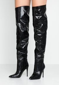 Botas negras sobre la rodilla con un patrón de cocodrilo texturizado y brillante, punta afilada y tacón de aguja alto. Parte superior suave con detalles fruncidos.