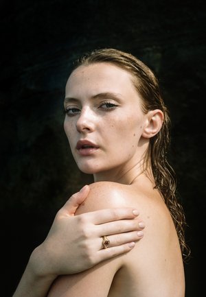 Ung kvinde med vådt hår kigger over skulderen, holder det med én hånd prydet med en guldring med en rød sten, mørk baggrund.