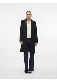 Abrigo largo negro con solapas en corte de muesca y dos botones frontales sobre una blusa clara de escote en v, combinado con jeans de pierna ancha oscuros y zapatos negros.