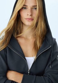 Felpa con zip di colore grigio scuro, dotata di cappuccio, con una texture liscia e zip in argento. Indossata sopra un top bianco, con i capelli che coprono parzialmente il viso.