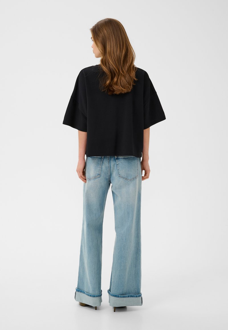 Femme aux cheveux bruns mi-longs portant un t-shirt noir ample et un jean large bleu clair avec revers, debout de dos.