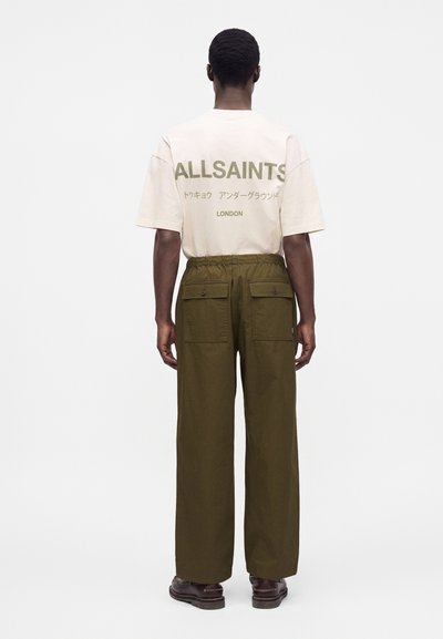 Άνδρας από πίσω, φοράει λευκό t-shirt AllSaints με κείμενο και χακί φαρδιά παντελόνια με πίσω τσέπες με καπάκι.
