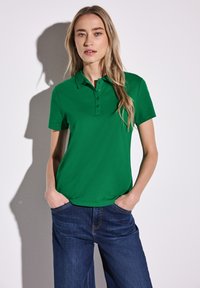 Grön pikétröja med treknappsstängning och korta ärmar, tillsammans med blå denimbyxor. Enkelt design med en mjuk textur.