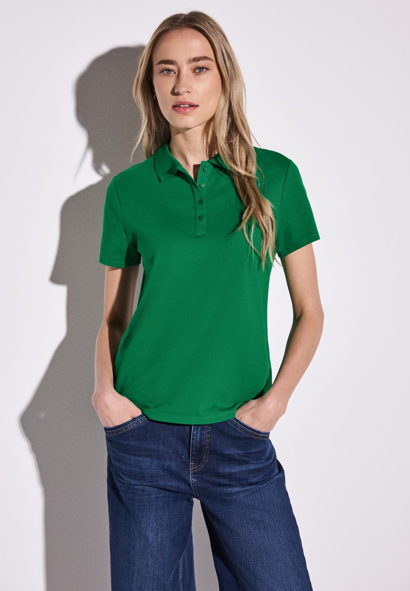 Grön pikétröja med treknappsstängning och korta ärmar, tillsammans med blå denimbyxor. Enkelt design med en mjuk textur.