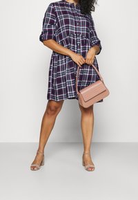 Lila kariertes Kleid mit einem Button-Down-Kragen und geraffter Taille, kombiniert mit einer rosa Handtasche aus Kunstleder und beigen Blockabsatzsandalen.