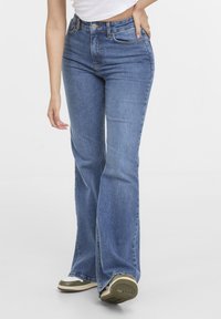 Jean bleu coupe slim avec légère évasement, porté avec des baskets blanches et un top blanc court, main posée sur la hanche.