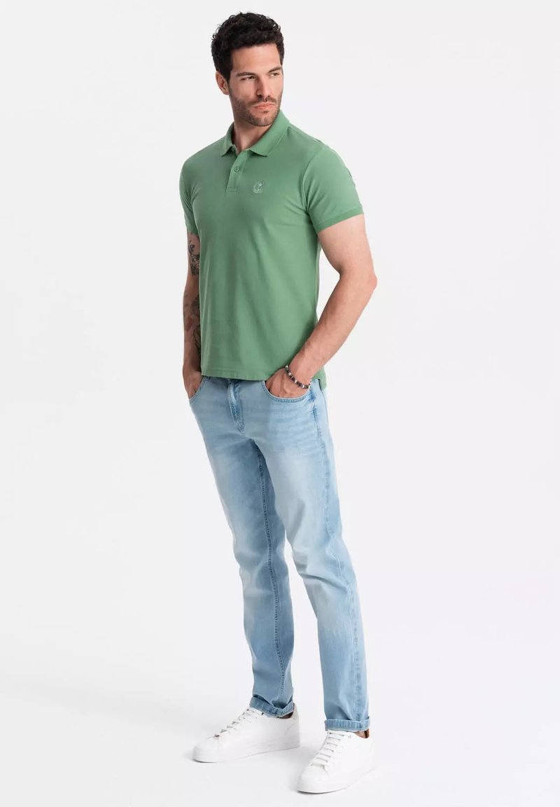 Polo de algodón verde con mangas cortas, combinado con jeans azul claro y zapatillas blancas. Presenta un logotipo en el lado izquierdo del pecho.