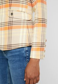 Osoba w pomarańczowo-beżowej marynarce w kratę z złotymi guzikami i niebieskimi dżinsami, pokazująca szczegóły kieszeni bocznej i rękawów.