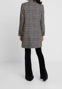 Grå rutig kappa med skräddarsydd passform, utrustad med hög krage och långa ärmar. Matchas med svarta utsvängda byxor för en strömlinjeformad silhuett.