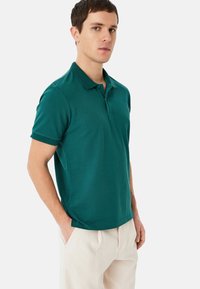 Teal polo skjorta med krage och korta ärmar, tillverkad av strukturerat tyg. Bärs med ljusa byxor.
