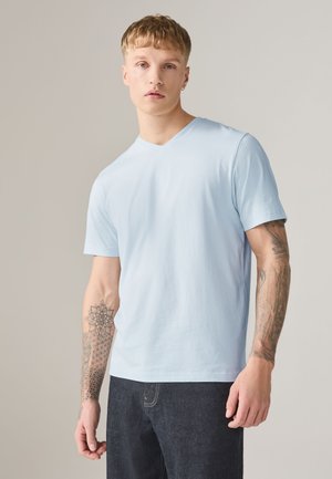 Młody mężczyzna w jasnoniebieskiej koszulce z dekoltem w serek i ciemnych dżinsach, pokazujący szczegółowe geometryczne i kwiatowe tatuaże na obu przedramionach.