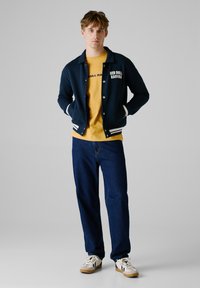 Marineblå bomberjakke med hvide striber, gul skjorte med trykt tekst, blå jeans og cremefarvede sneakers med sorte detaljer.