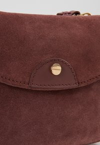 Brązowa zamszowa torba z zaokrągloną klapą, wyposażona w złote zapięcie na zatrzask. Gładkie skórzane akcenty i widoczne detale stębnowania.