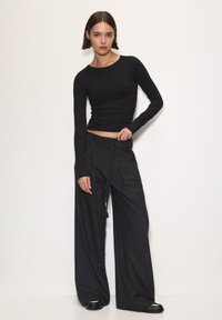 Egy nő áll a fehér falnak támaszkodva, testhezálló fekete hosszúujjú felsőt, széles szárú csíkos nadrágot és fekete cipőt visel.