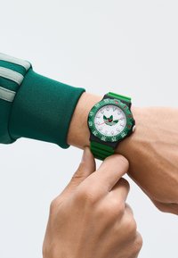 Roheline käekell valge kellaga ja roheliste detailidega, kummist rihm, must bezel minutimarkeritega, keskosas logo.