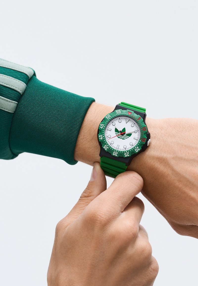 Roheline käekell valge kellaga ja roheliste detailidega, kummist rihm, must bezel minutimarkeritega, keskosas logo.