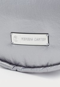 Sac rembourré argenté avec une surface texturée et une finition brillante. Comprend une étiquette en métal rectangulaire avec "VENEDA CARTER" gravé.