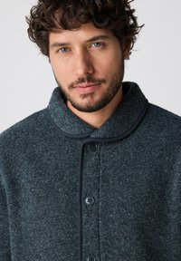 Giacca di lana grigio scuro con chiusura a bottoni, texture morbida e colletto alto. Presenta piping nero a contrasto lungo i bordi.