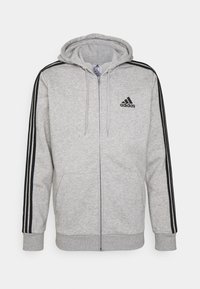 Sudadera gris con cremallera, con rayas negras en los hombros, puños acanalados y capucha. Presenta un logo negro de Adidas bordado en el pecho.