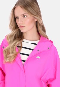 Világos rózsaszín kapucnis esőkabát, amelyen a mellkason világosszürke esernyő logó és ezüst patentzárak láthatók. Csíkos ing alatta.