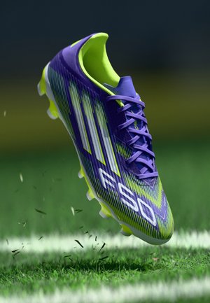 Botte de football violette et verte avec une surface texturée, trois bandes blanches, et un design léger, possédant des crampons multidirectionnels et des lacets.