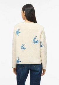 Ženska z dolgimi temnimi lasmi, oblečena v kremno jakno iz flisa z modrimi cvetličnimi vzorci in temno modre kavbojke, obrnjena stran na beli podlagi.