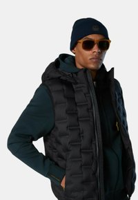 Chaleco puffer negro con patrón acolchado, llevado sobre una sudadera con capucha verde oscura. Los accesorios incluyen un gorro de punto azul marino y gafas de sol marrones.