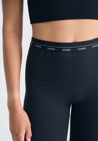 Juodos legginsai, pagaminti iš lygaus, elastingo medžiagos su GUESS prekės ženklo elastine juosta. Prigludusi forma pabrėžia liemens liniją.