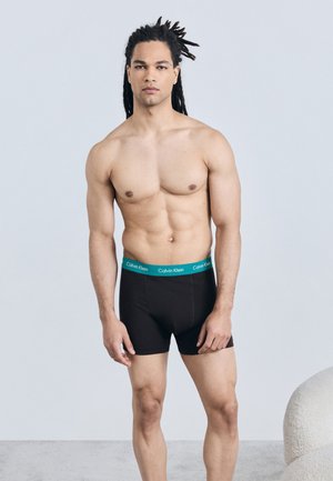 Mann mit Dreadlocks, der schwarze Calvin Klein Boxershorts trägt und vor einem schlichten, hellen Hintergrund steht.