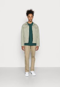 Jaqueta de denim verde-claro, sweatshirt verde-escuro, calças bege e ténis brancos. Design simples com botões e bolsos na jaqueta.