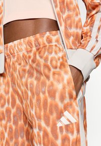 Oranži rakstainas brīvā stila bikses ar leoparda rakstu, izgatavotas no gludas auduma, ar baltu Adidas logotipu un kontrastējošām baltām svītrām.