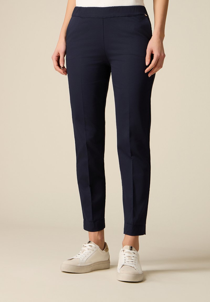 Femeie purtând pantaloni bleumarin slim până la gleznă și adidași albi cu detalii leopard pe toc, stând în fața unui fundal simplu.