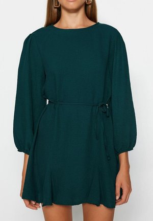 Vrouw draagt een donkergroene mini-jurk met lange mouwen, een dunne riem in de taille geknoopt en pofmouwen die eindigen in elastische manchetten.