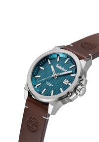 Reloj de acero inoxidable con esfera texturizada azul, marcadores plateados y correa de cuero marrón. Incluye una pantalla de fecha y detalles en la corona.
