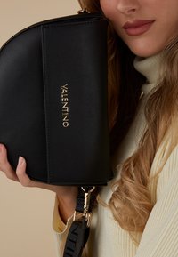 Sac noir en cuir avec un design incurvé, arborant un logo doré "Valentino", une bandoulière détachable et des accents en matériel doré.