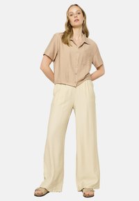 Light brown short-sleeve blouse with a button front and pocket, paired with loose-fitting, cream wide-leg trousers and beige sandals.